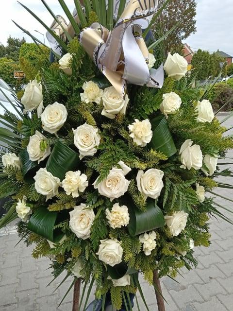 Duży wieniec pogrzebowy z białych róż i goździków, z zielenią i złoto-białą kokardą, na stojaku. 