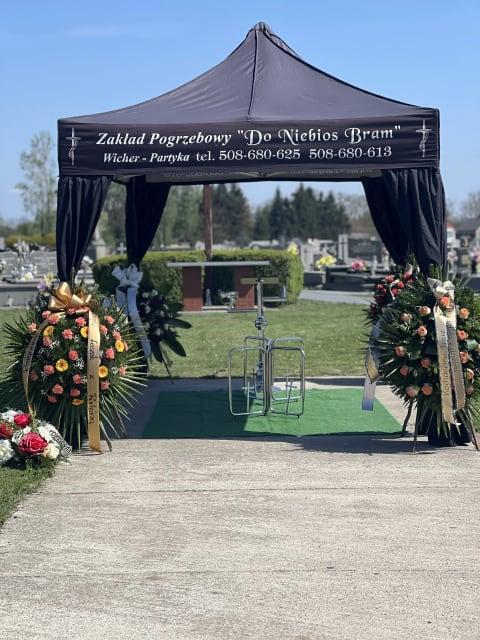 Czarny namiot pogrzebowy z napisami "Zakład Pogrzebowy" oraz "Do Niebios Bram", ustawiony na cmentarzu, otoczony dużymi wieńcami kwiatowymi.